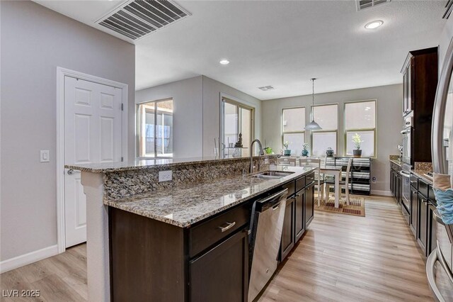 11036 Mount Hunter St, Las Vegas, NV 89179 - kitchen featuring appliances with stainless steel finishes, light wood-type flooring, dark brown cabinetry, a center island with sink, and decorative light fixtures