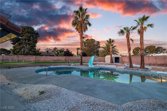 pool at dusk with an outdoor fireplace, a fenced backyard, a water slide, a patio, and a fenced in pool