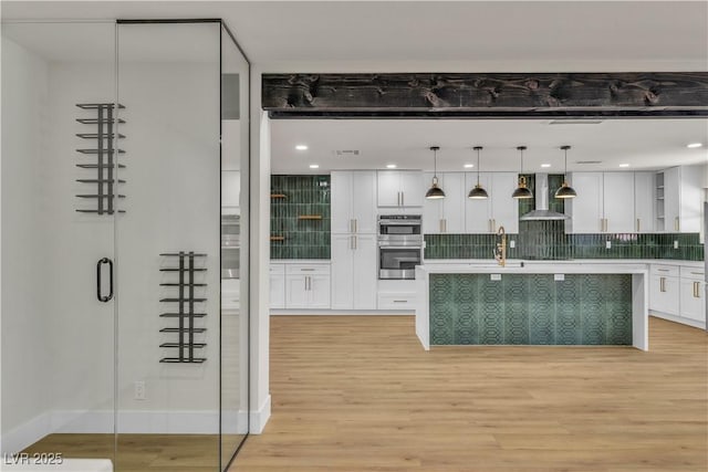 kitchen featuring double oven, wall chimney range hood, white cabinetry, light countertops, and light wood finished floors