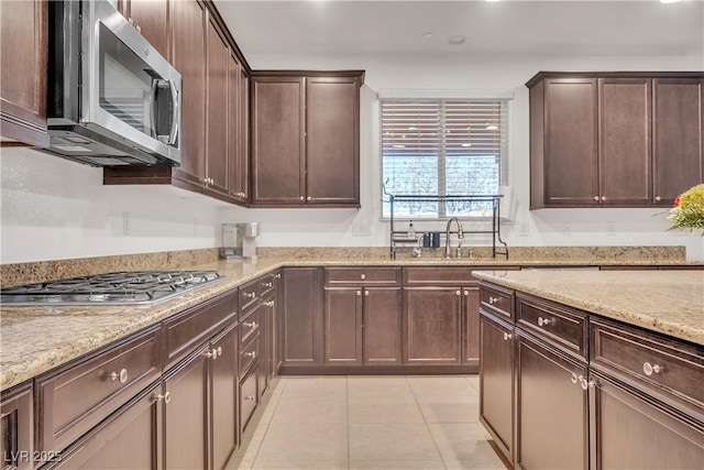 kitchen with light tile patterned floors, a sink, dark brown cabinets, light stone countertops, and stainless steel appliances