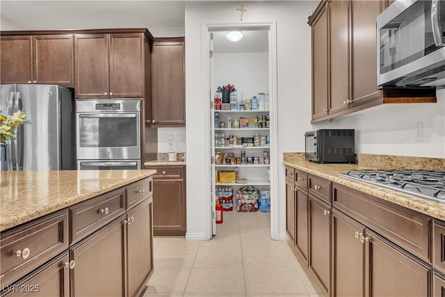 kitchen with dark brown cabinetry, a toaster, light tile patterned floors, light stone countertops, and stainless steel appliances