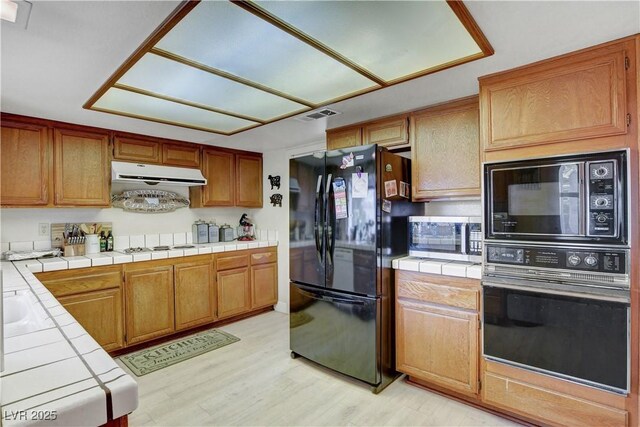 kitchen with tile countertops, light wood-type flooring, visible vents, black appliances, and under cabinet range hood