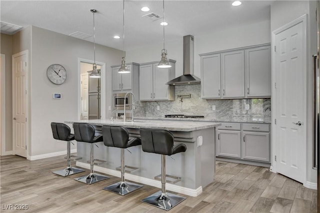 kitchen with light wood-style floors, wall chimney range hood, gray cabinets, light stone counters, and backsplash