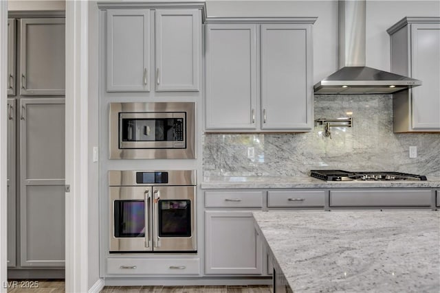 kitchen with wall chimney range hood, appliances with stainless steel finishes, gray cabinetry, and light stone countertops