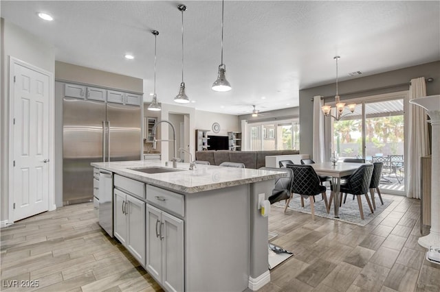 kitchen with light stone counters, light wood-style flooring, a center island with sink, appliances with stainless steel finishes, and pendant lighting