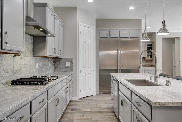 kitchen featuring wall chimney exhaust hood, a sink, an island with sink, stainless steel appliances, and gray cabinets
