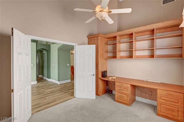unfurnished office with light carpet, built in study area, visible vents, ceiling fan, and arched walkways