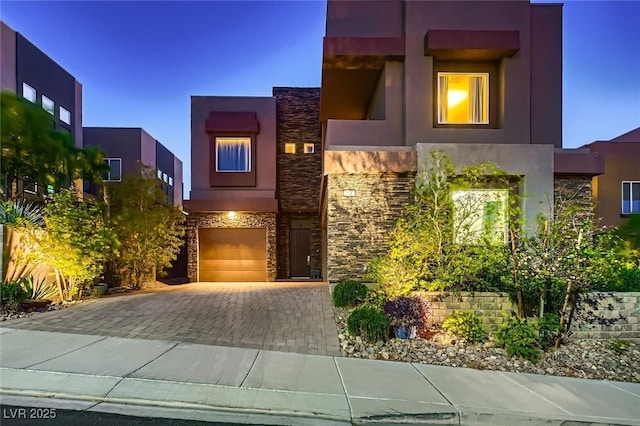 contemporary house with stone siding, an attached garage, stucco siding, and decorative driveway