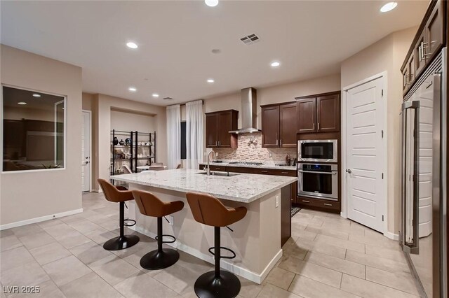 kitchen featuring a sink, backsplash, built in appliances, wall chimney range hood, and an island with sink