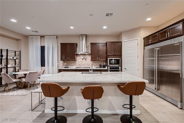 kitchen with wall chimney range hood, dark brown cabinets, a sink, built in appliances, and visible vents