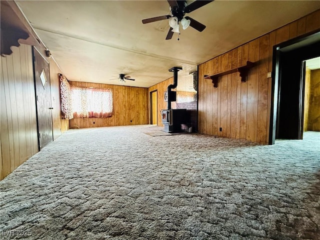 unfurnished living room with wood walls, a wood stove, a ceiling fan, and carpet flooring