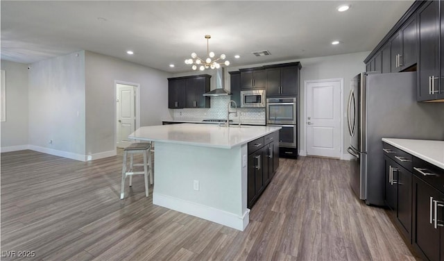 kitchen featuring wall chimney exhaust hood, visible vents, a kitchen island with sink, stainless steel appliances, and a notable chandelier