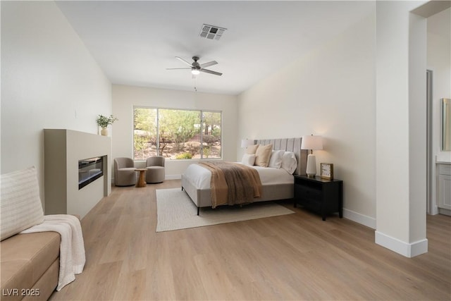 bedroom featuring a glass covered fireplace, light wood-style flooring, baseboards, and ceiling fan