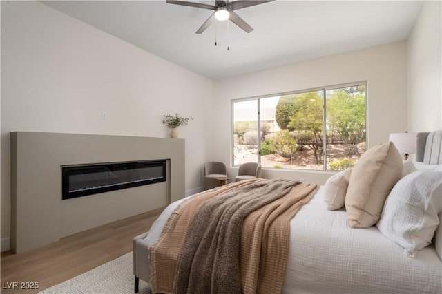 bedroom featuring wood finished floors, a glass covered fireplace, and a ceiling fan