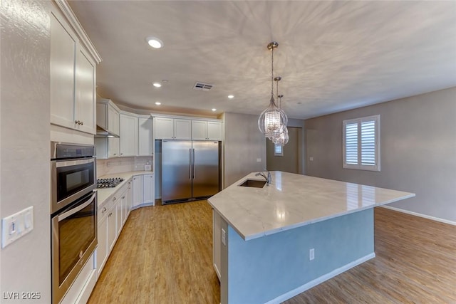 kitchen with a sink, light wood finished floors, a center island with sink, and appliances with stainless steel finishes