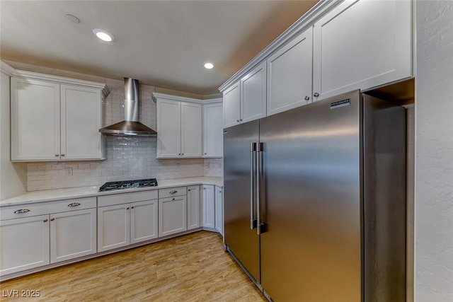 kitchen with wall chimney range hood, tasteful backsplash, appliances with stainless steel finishes, light countertops, and light wood-style floors