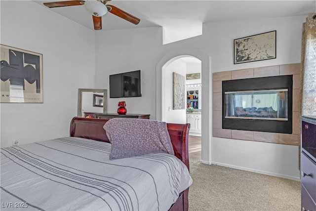 carpeted bedroom with arched walkways, a tiled fireplace, ensuite bath, and a ceiling fan