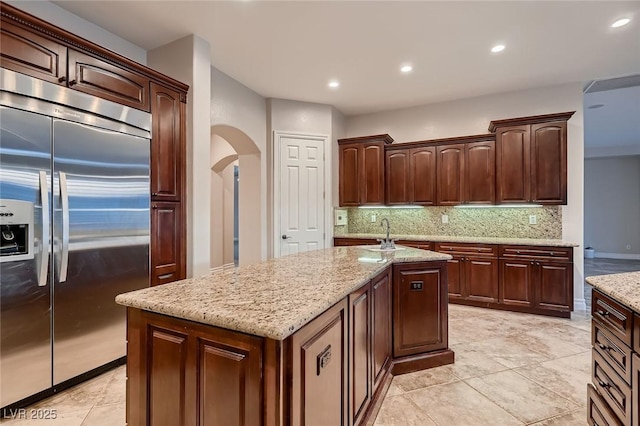 kitchen featuring tasteful backsplash, stainless steel built in refrigerator, light stone countertops, a kitchen island with sink, and arched walkways