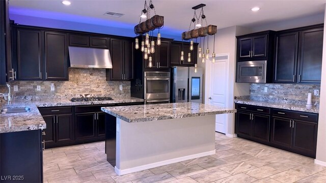 kitchen with appliances with stainless steel finishes, under cabinet range hood, a sink, tasteful backsplash, and light stone countertops