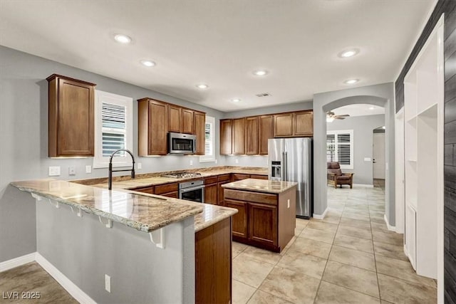 7806 Sundial Peak St, Las Vegas, NV 89166 - kitchen featuring appliances with stainless steel finishes, a peninsula, light stone counters, brown cabinets, and recessed lighting
