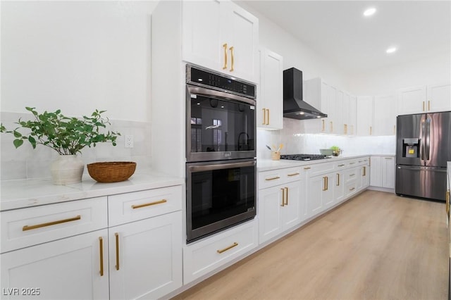 kitchen with tasteful backsplash, light wood-type flooring, wall chimney range hood, white cabinets, and stainless steel appliances