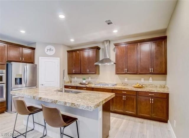 kitchen featuring wall chimney exhaust hood, a kitchen breakfast bar, a kitchen island with sink, a sink, and stainless steel fridge