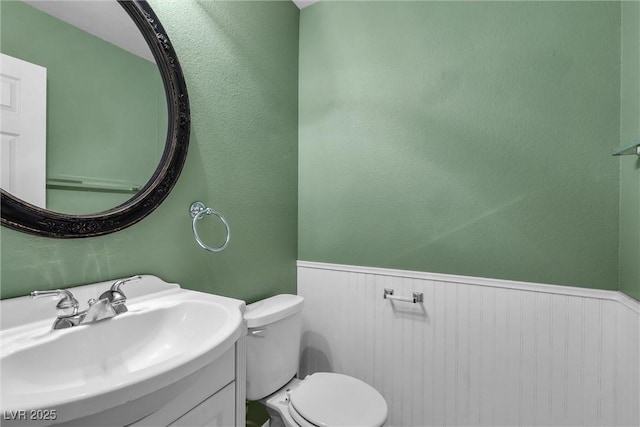bathroom featuring a textured wall, toilet, vanity, and a wainscoted wall