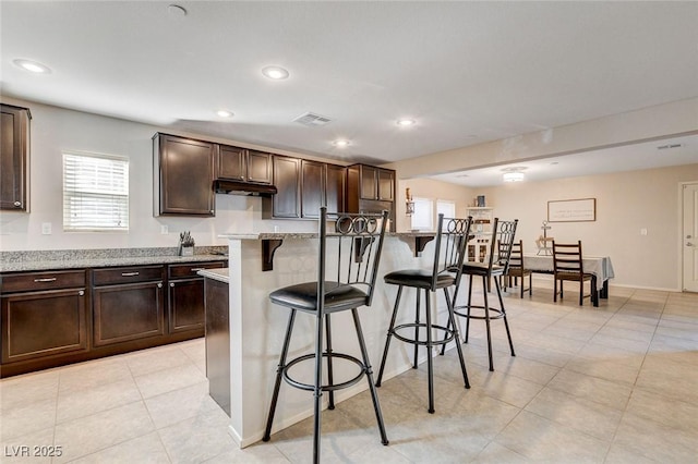 kitchen featuring visible vents, light tile patterned floors, a kitchen bar, under cabinet range hood, and dark brown cabinets