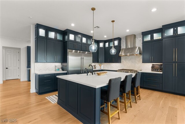 kitchen featuring appliances with stainless steel finishes, wall chimney range hood, light countertops, light wood-style flooring, and tasteful backsplash