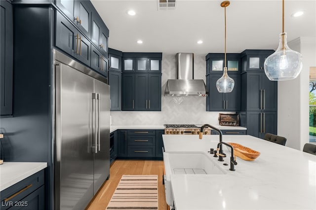 kitchen with wall chimney range hood, stainless steel built in refrigerator, a sink, light countertops, and light wood-style floors