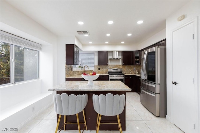 kitchen with a breakfast bar, wall chimney range hood, dark brown cabinets, visible vents, and stainless steel appliances