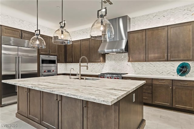 kitchen with wall chimney exhaust hood, dark brown cabinetry, appliances with stainless steel finishes, a sink, and decorative backsplash