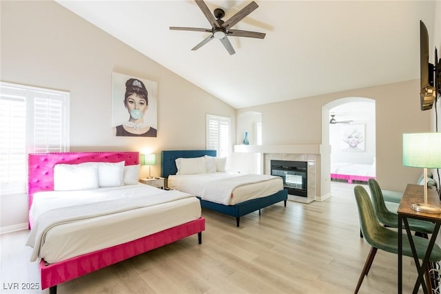 bedroom with baseboards, arched walkways, a tiled fireplace, light wood finished floors, and vaulted ceiling