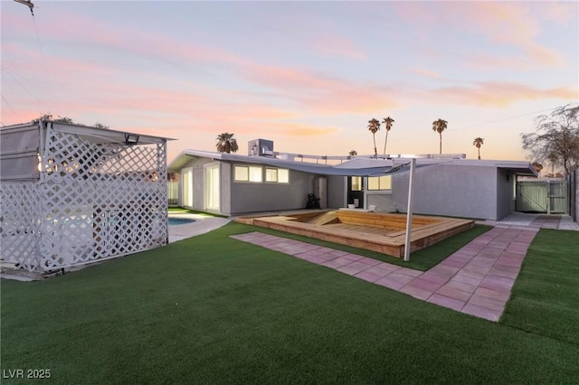 back of property at dusk with fence, a gate, a lawn, and stucco siding