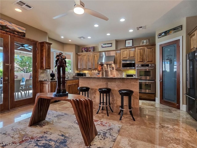 kitchen with ceiling fan, stainless steel appliances, wall chimney range hood, decorative backsplash, and a center island