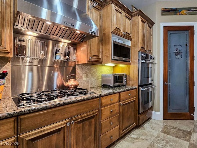 kitchen featuring stainless steel appliances, exhaust hood, backsplash, and dark stone countertops