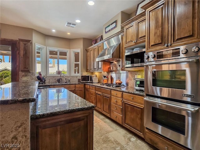 kitchen with stainless steel appliances, wall chimney exhaust hood, decorative backsplash, dark stone countertops, and a sink