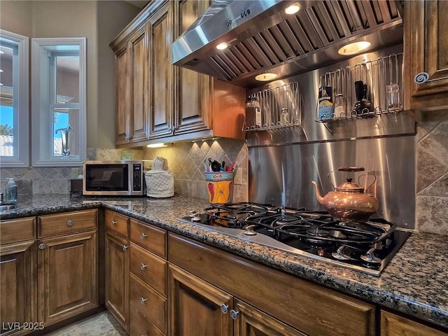kitchen featuring wall chimney range hood, black gas cooktop, dark stone countertops, decorative backsplash, and stainless steel microwave