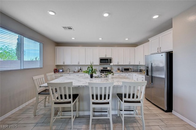 kitchen with light stone counters, a center island with sink, white cabinets, appliances with stainless steel finishes, and visible vents