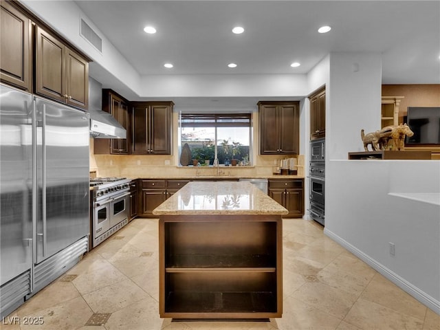 kitchen featuring backsplash, wall chimney range hood, a sink, light stone countertops, and built in appliances