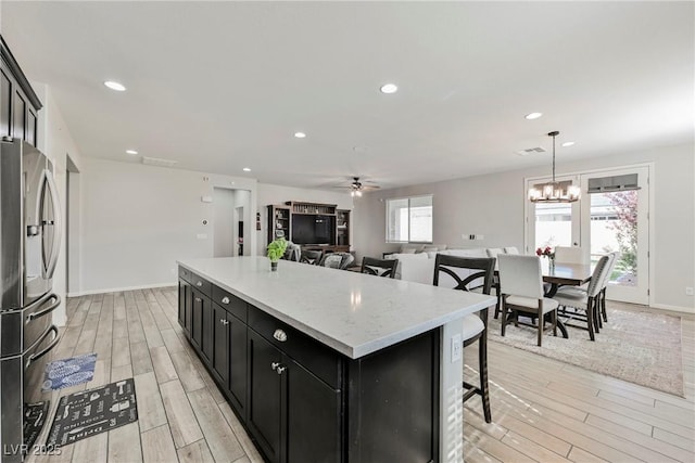 kitchen featuring a kitchen island, light wood-type flooring, stainless steel fridge, ceiling fan with notable chandelier, and dark cabinets