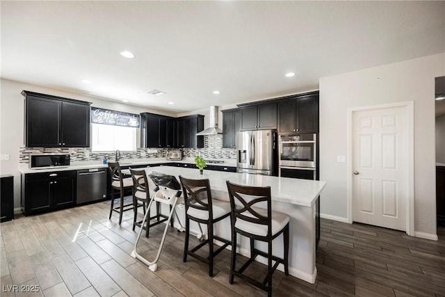 kitchen featuring dark cabinetry, stainless steel appliances, wall chimney exhaust hood, and a kitchen breakfast bar