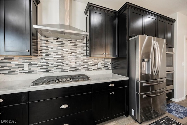 kitchen featuring wall chimney range hood, stainless steel appliances, dark cabinetry, and tasteful backsplash