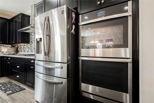 kitchen featuring light wood-type flooring, wall chimney exhaust hood, dark cabinetry, backsplash, and stainless steel appliances