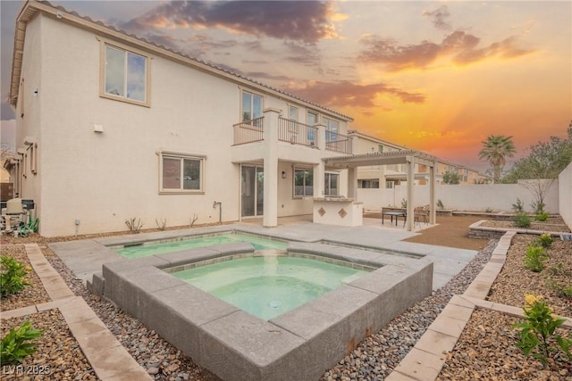 back of property at dusk with a patio area, a balcony, a fenced backyard, an in ground hot tub, and stucco siding