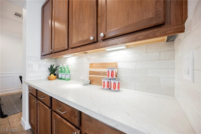 kitchen featuring visible vents, backsplash, brown cabinetry, light countertops, and ornamental molding