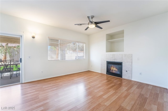 unfurnished living room featuring built in shelves, ceiling fan, a tile fireplace, and light wood-type flooring