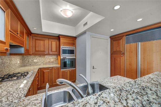 kitchen featuring light stone countertops, a sink, built in appliances, brown cabinetry, and visible vents
