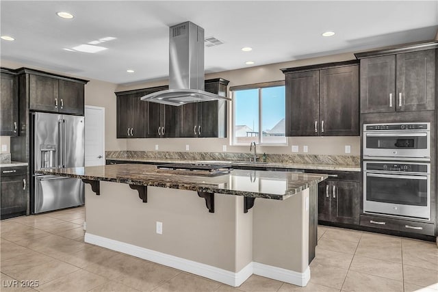 kitchen featuring stainless steel appliances, island exhaust hood, dark stone countertops, a breakfast bar area, and a sink