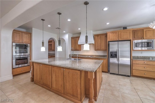 kitchen with wall chimney range hood, light tile patterned floors, brown cabinets, appliances with stainless steel finishes, and arched walkways